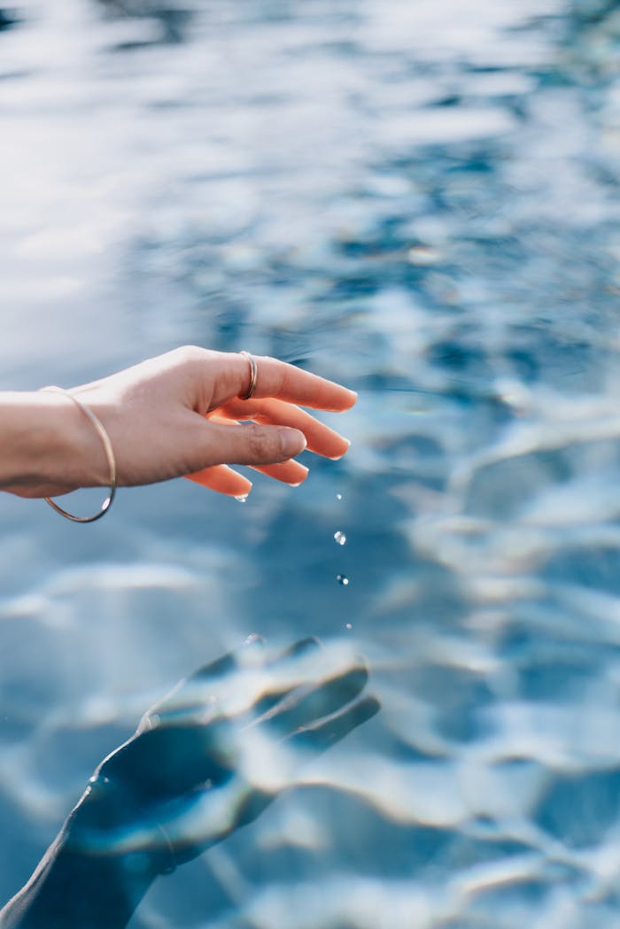 A woman's hand gently touches the clear blue water surface creating serene ripples.