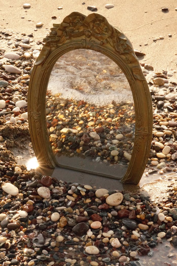 A vintage mirror reflects the vibrant pebble beach, creating an artistic and surreal shoreline scene.